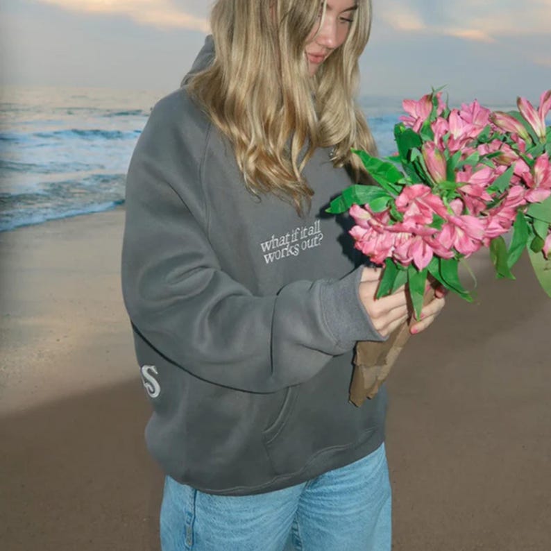 May include: A gray hoodie with the text "what if it all works out?" and a white "S" on the sleeve. The person is holding a bouquet of pink flowers. The background shows a beach and ocean.