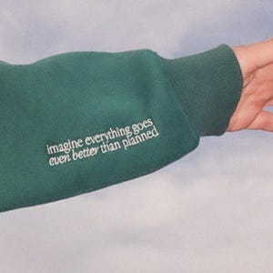 May include: A dark green sweatshirt sleeve with the embroidered text "imagine everything goes even better than planned" in white. The sleeve is ribbed at the cuff. The background is a light blue.