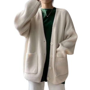 May include: An oversized cream-colored knit cardigan featuring a V-neckline, long sleeves with ribbed cuffs, and a button-down front closure. The garment includes two large patch pockets on the front and is layered over a dark green shirt.