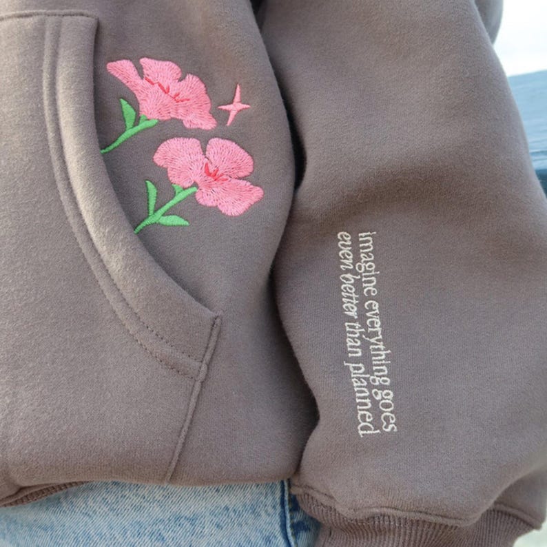 May include: A brown hoodie with embroidered pink flowers and a star on the front pocket. The sleeve features the text "imagine everything goes even better than planned" in white.