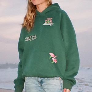 May include: A dark green hoodie with a front pocket and a hood. The hoodie features floral embroidery and the text "darling" above the pocket and "what if it all works out?" on the left sleeve.