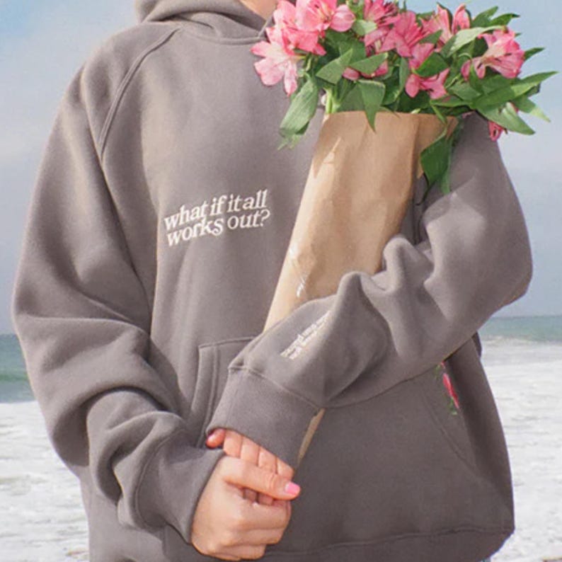 May include: A gray hoodie with the text "what if it all works out?" on the chest. The hoodie has a front pocket and long sleeves. The person is holding a bouquet of pink flowers in a brown paper bag.