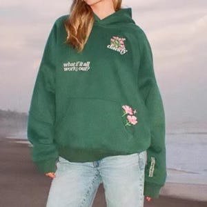 May include: A dark green hoodie with a front pocket and a hood. The hoodie has the text "what if it all works out?" and "dandy" with pink floral embroidery. The model is wearing light blue jeans.