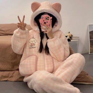 Könnte beinhalten: Hellrosa, flauschiges Pyjama-Set mit Kapuze und Bärenohren. Der Pyjama hat ein Karomuster und eine Cartoon-Hunde-Stickerei auf der Brust. Das Set beinhaltet ein Oberteil mit Reißverschluss und eine passende Hose, die für Komfort und Wärme konzipiert sind.