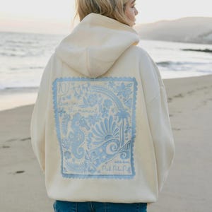 May include: Cream-colored hoodie with a light blue graphic on the back. The graphic features a seahorse, flowers, and the text "Live in the moment" and "Pink Palm Puff". The hoodie has a hood and a relaxed fit.