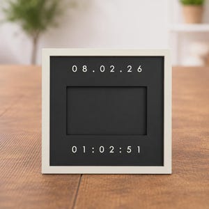 Op de afbeelding: Een vierkant, wit omrand zwart letterbord toont de datum en tijd: 08.02.26 en 01:02:51. Het bord heeft een rechthoekige uitsparing in het midden. Het bord staat op een houten oppervlak.