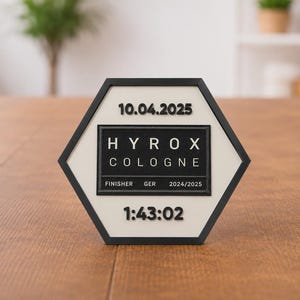 Op de afbeelding: Een zwart-wit zeshoekige plaquette met de tekst "10.04.2025", "HYROX COLOGNE", "FINISHER GER 2024/2025" en "1:43:02". De plaquette ligt op een houten oppervlak.