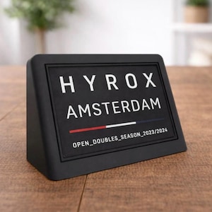 以下が含まれることがあります： 黒い長方形のサインで、白い文字で「HYROX AMSTERDAM」と書かれています。テキストの下には、赤と白のセクションがある水平線があります。サインには「OPEN_DOUBLES_SEASON_2023/2024」も含まれています。