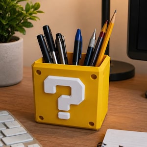 May include: A yellow cube-shaped pencil holder featuring a raised white question mark on the front face. The container has four circular indentations at the corners and is filled with various pens, pencils, and a mechanical pencil for desk storage.