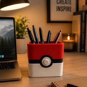 May include: A square desk organiser designed to resemble a red, white, and black Poké Ball. The container features a central circular button detail on the front and holds several pens and pencils upright within its open top compartment.