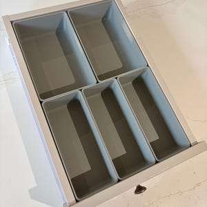 May include: A light-coloured wooden drawer organiser featuring five rectangular compartments with smooth, light grey plastic inserts. The organiser is designed to fit inside a drawer, providing structured storage space for various small items.