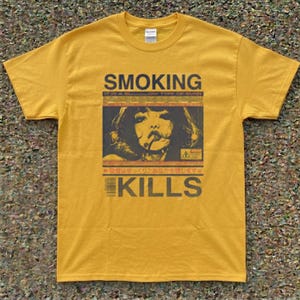 Op de afbeelding: Een mosterdgeel T-shirt met korte mouwen en een grafische print met de tekst SMOKING KILLS. Het ontwerp bevat een centrale illustratie van een rokend persoon, een streepjescode en Japanse karakters, alles uitgevoerd in donkerbruin.