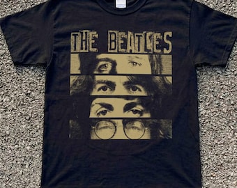 Könnte beinhalten: Ein schwarzes Kurzarm-T-Shirt mit dem Schriftzug THE BEATLES in einer abgenutzten goldenen Schrift über einer Grafik aus vier Augenpaaren in horizontalen Feldern, wobei das untere Augenpaar eine runde Brille trägt, alles in Gold gedruckt.