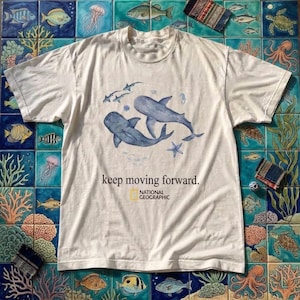 Op de afbeelding: Een crèmekleurig T-shirt met korte mouwen en een blauwe aquarelillustratie van twee walvissen, kleine vissen en een zeester. De tekst "keep moving forward." is in het zwart gedrukt, gevolgd door het geel-witte National Geographic logo.