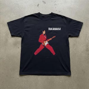Könnte beinhalten: Ein schwarzes Kurzarm-T-Shirt mit einer Grafik einer Person im roten Anzug, die eine E-Gitarre spielt. Der Text TAKANAKA ist in weißen Großbuchstaben über der Grafik aufgedruckt. Das Shirt hat einen Rundhalsausschnitt und einen geraden Saum.