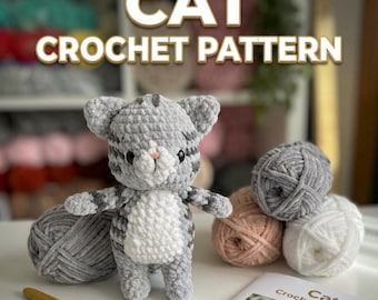 May include: A gray and white crocheted cat plush toy with dark stripes, black eyes, and a pink nose. The image includes text reading CAT CROCHET PATTERN, several balls of yarn in gray, white, and peach, and a yellow crochet hook on a surface.