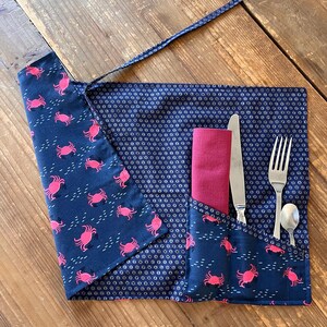 Peut inclure: Porte-ustensiles en tissu bleu marine avec un motif de crabes roses. Il contient une serviette rouge, un couteau, une fourchette et une cuillère. Conçu pour être enroulé et maintenu par une attache.