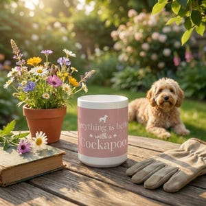 Op de afbeelding: Een witte keramische mok met een roze band met de tekst "Everything is better with a Cockapoo" en een honden silhouet. De mok staat op een houten tafel met een boek, bloemen en tuinhandschoenen. Een Cockapoo hond is op de achtergrond.