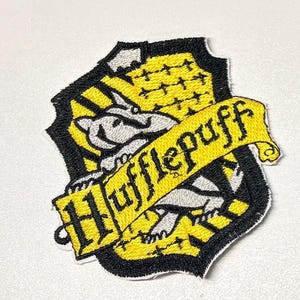 Może przedstawiać: Haftowana naszywka z herbem Hufflepuff, przedstawiająca borsuka, w kolorach żółtym i czarnym, z napisem "Hufflepuff" na żółtym banerze. Naszywka ma kształt tarczy z czarną obwódką.