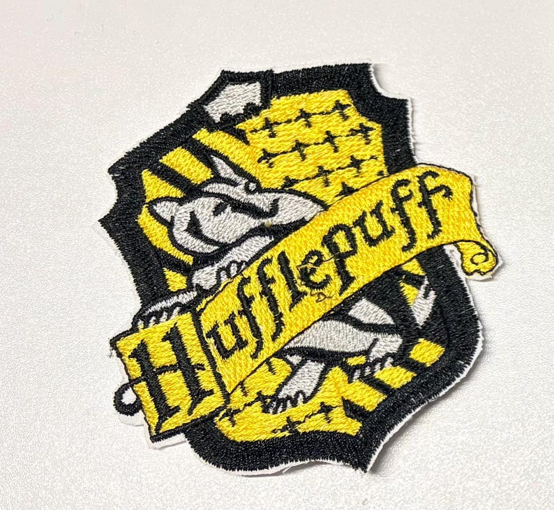 Pode incluir: Patch bordado do bras&atilde;o da Hufflepuff, com um texugo, cores amarelo e preto, e a palavra "Hufflepuff" em uma faixa amarela. O patch tem formato de escudo com uma borda preta.
