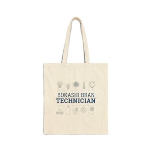 May include: A cream-colored tote bag with the text "BOKASHI BRAN TECHNICIAN" in navy blue. The bag features illustrations of plants and gardening tools above and below the text. The tote bag has two cream-colored handles.