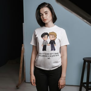 May include: White t-shirt featuring a cartoon illustration of two figures holding hands. The text on the shirt reads "HOLY F*CKING SH*T. SHANE HOLLANDER IS IN LOVE WITH ME."