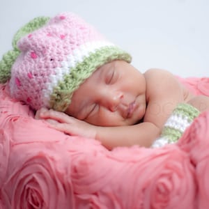 May include: A sleeping baby wearing a pink and green crocheted hat and matching wrist cuffs, lying on a pink rose-shaped blanket.
