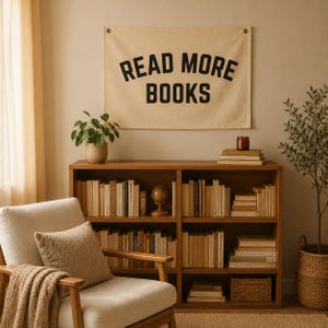 May include: A rectangular cream-colored fabric wall hanging featuring the black text READ MORE BOOKS. The banner is mounted above a wooden bookshelf filled with various books, a small globe, and a woven basket, positioned near a beige armchair.