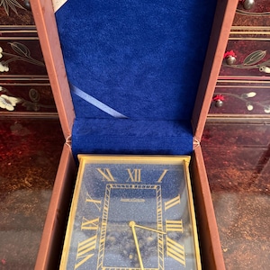 May include: A rectangular, gold-toned clock with a dark blue face and gold Roman numerals. The clock is in a brown box with a blue velvet interior. The brand name "Jaeger LeCoultre" is visible on the clock face.