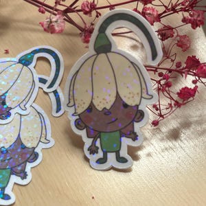 May include: A holographic sticker featuring a cartoon character with a pale yellow flower-shaped hat, a dark brown face, and a green shirt. The character has a curved stem extending from the top of the flower hat and a subtle glittery finish.