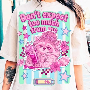 Puede incluir: Camiseta color crema con un mapache rosa con una bebida, rodeado de flores y estrellas rosas. La camiseta tiene el texto "Don't expect too much from me" en rosa y verde. También hay un gráfico de batería baja.