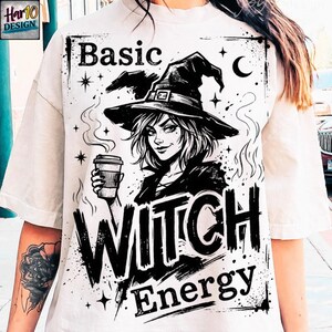 May include: White t-shirt with a black and white graphic of a witch wearing a hat and holding a coffee cup. The text on the shirt reads "Basic Witch Energy."