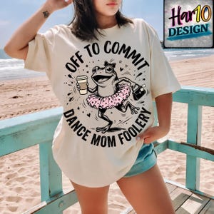 May include: Cream-colored t-shirt featuring a whimsical frog design. The frog is wearing a pink tutu and holding a coffee cup and purse. The text on the shirt reads "Off to Commit Dance Mom Foolery".