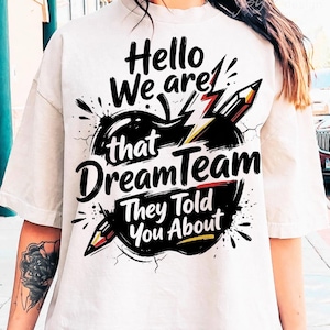 Puede incluir: Una camiseta blanca con un diseño gráfico negro que incluye el texto "Hello We are that DreamTeam They Told You About". El diseño presenta una gran forma de manzana negra, un rayo amarillo y dos lápices con detalles en rojo y amarillo.