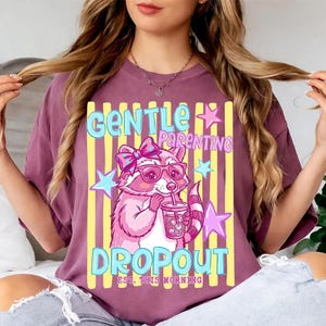 Puede incluir: Una camiseta color ciruela con un gráfico de un mapache rosa con un lazo y gafas, sosteniendo una bebida. El texto dice GENTLE PARENTING DROPOUT EST. THIS MORNING. El diseño incluye rayas verticales amarillas y estrellas de color azul claro.
