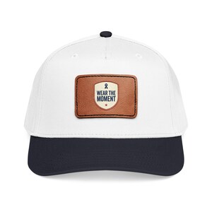 May include: A white baseball cap with a navy blue peak and a black button. It has a brown leather patch with the text "WEAR THE MOMENT" and a blue ribbon symbol.