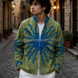 Pode incluir: Um casaco verde-azeitona de manga comprida com um design de aplicação azul vibrante em forma de sol na frente e nas mangas, gola mandarim e fecho de correr frontal. O texto ASSUHANDICRAFTINDIA está impresso no centro do casaco.