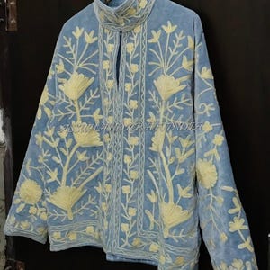 May include: A light blue jacket featuring a high collar and long sleeves, adorned with cream-colored floral embroidery throughout the front and sleeves. The text ASSUHANDICRAFTINDIA is visible across the center of the garment.