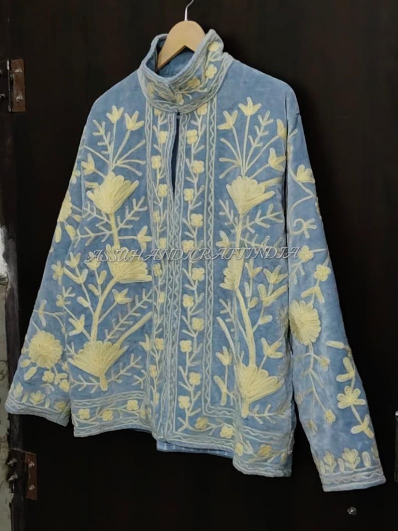 May include: A light blue jacket featuring a high collar and long sleeves, adorned with cream-colored floral embroidery throughout the front and sleeves. The text ASSUHANDICRAFTINDIA is visible across the center of the garment.