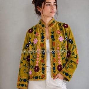 Op de afbeelding: Een olijfgroene jas met een opstaande kraag en lange mouwen, versierd met levendig bloemenborduursel in roze, donkerrode en blauwe tinten. Het kledingstuk wordt gedragen over een wit hemd en een witte broek, met de tekst ASSUHANDICRAFTINDIA zichtbaar.