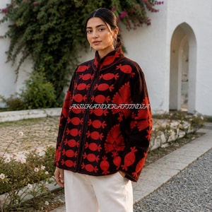 Può includere: Una giacca in velluto marrone scuro con collo alto, maniche lunghe e chiusura frontale con zip. La giacca è decorata con vivaci ricami rossi raffiguranti forme stilizzate di melograno e motivi floreali. Il testo ASSUHANDICRAFTINDIA è visibile.