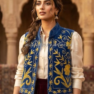 Op de afbeelding: Een blauw fluwelen vest met gele bloemenborduursels en een opstaande kraag, gedragen over een crèmekleurig overhemd met lange mouwen en een donkerrode fluwelen rok. De tekst ASSUHANDICRAFTINDIA is zichtbaar op de voorzijde van het vest.