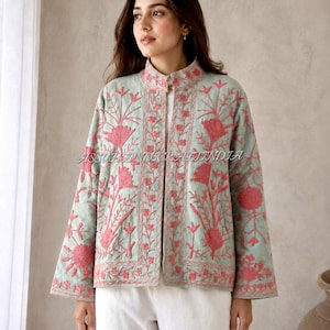 Könnte beinhalten: Eine hellgrüne, gesteppte Jacke mit Stehkragen und langen Ärmeln, verziert mit aufwendigen rosa Blumenstickereien auf der Vorderseite und den Ärmeln. Das Kleidungsstück ist vorne offen und enthält den Text ASSUHANDICRAFTINDIA.