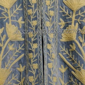 May include: A light blue jacket featuring a high collar and intricate yellow floral embroidery throughout the front panels. The garment displays vertical borders with small flower patterns and the text ASSUHANDICRAFTINDIA.