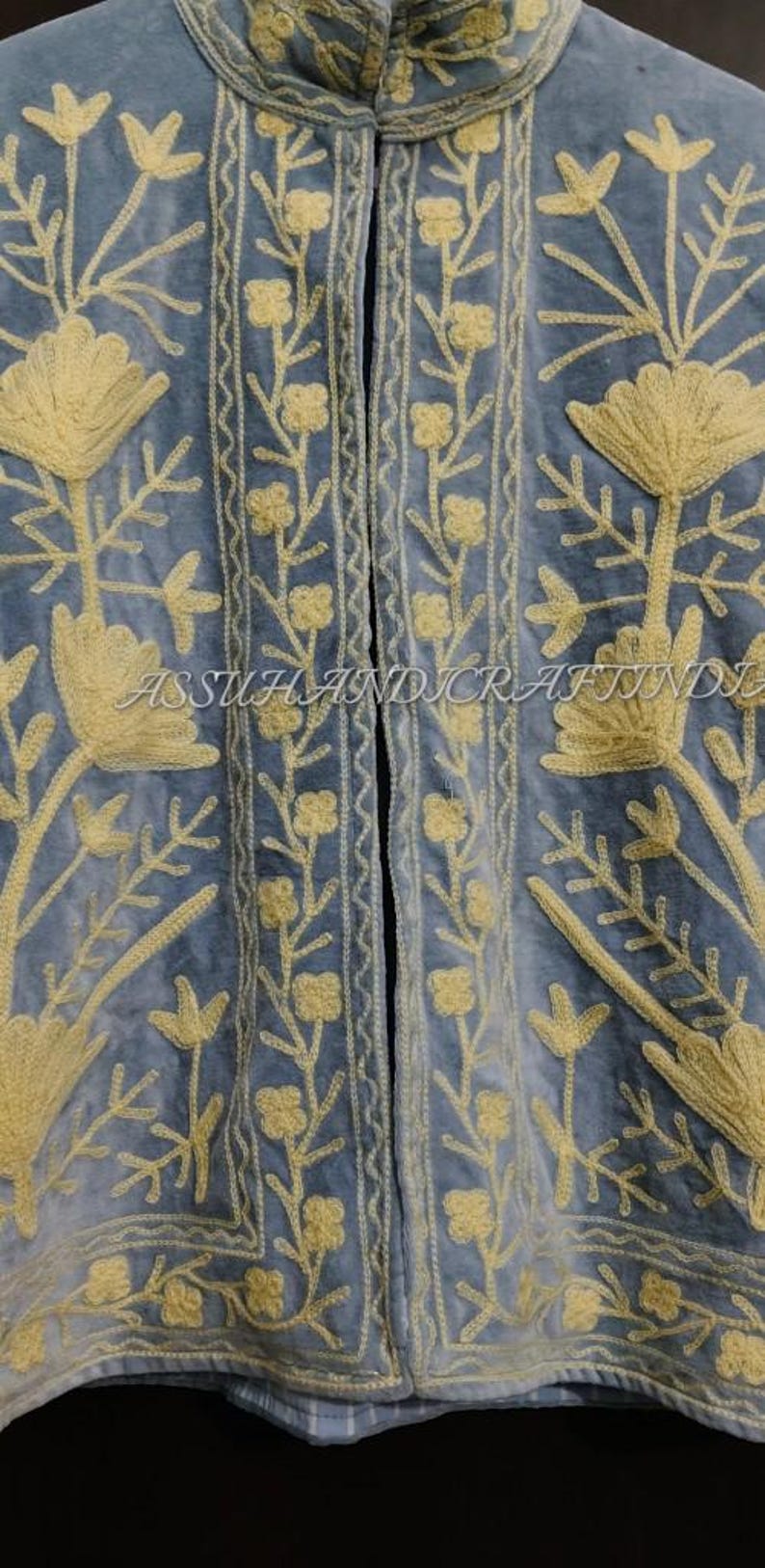 May include: A light blue jacket featuring a high collar and intricate yellow floral embroidery throughout the front panels. The garment displays vertical borders with small flower patterns and the text ASSUHANDICRAFTINDIA.