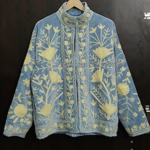 May include: A light blue jacket featuring a high collar and long sleeves, decorated with cream-colored floral embroidery throughout the front and sleeves. The text ASSUHANDICRAFTINDIA is visible in the center of the garment.