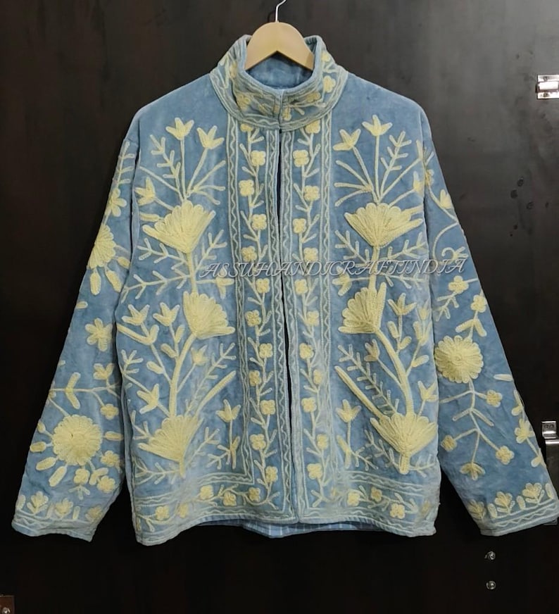 May include: A light blue jacket featuring a high collar and long sleeves, decorated with cream-colored floral embroidery throughout the front and sleeves. The text ASSUHANDICRAFTINDIA is visible in the center of the garment.