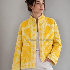 Könnte beinhalten: Eine leuchtend gelbe Steppjacke mit aufwendiger, tonaler Blumenstickerei auf dem gesamten Stoff. Die Jacke hat einen hohen Kragen, lange Ärmel und einen geraden Saum. Der Text ASSUHANDCRAFTINDIA ist in der vorderen Mitte zu sehen.