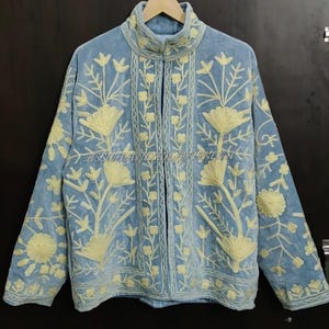May include: A light blue jacket featuring cream-colored floral embroidery throughout the front and sleeves. The garment has a high collar, long sleeves, and a central opening. The text ASSUHANDICRAFTINDIA is visible across the center.