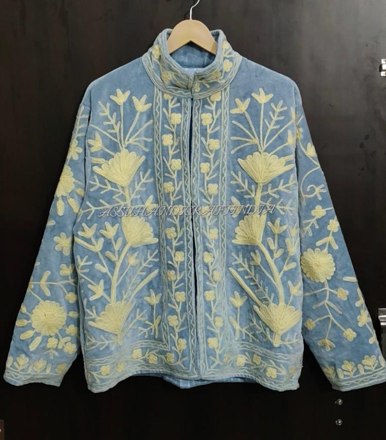 May include: A light blue jacket featuring cream-colored floral embroidery throughout the front and sleeves. The garment has a high collar, long sleeves, and a central opening. The text ASSUHANDICRAFTINDIA is visible across the center.